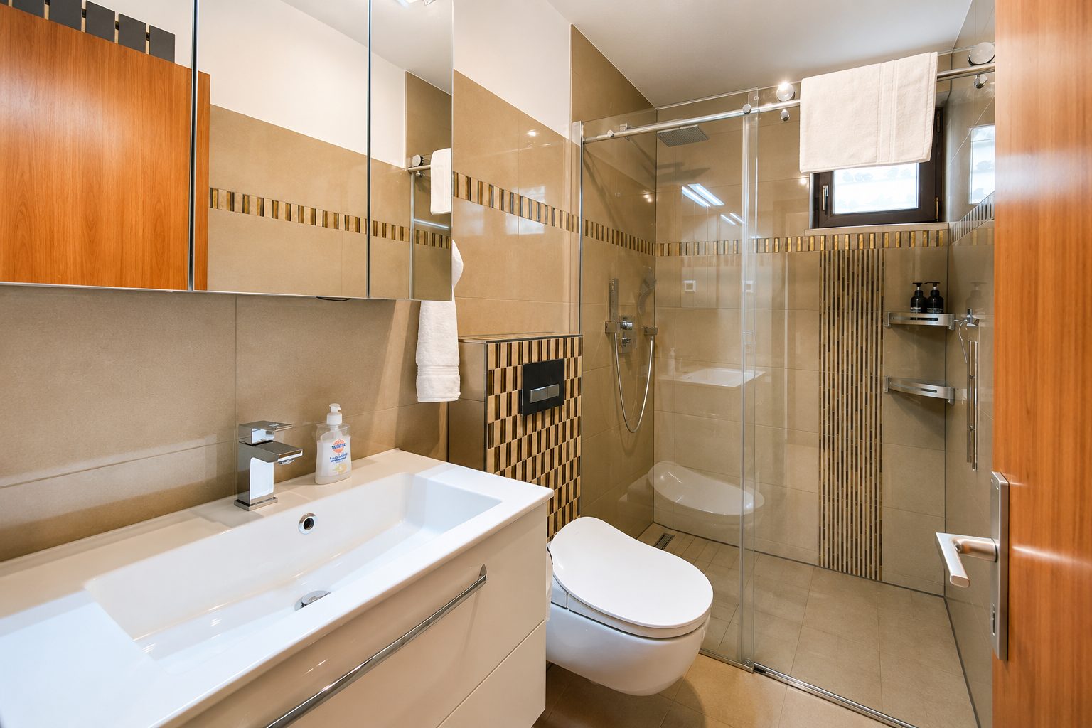 Badezimmer in Holz- und Beigetoenen mit Waschtisch, Wand-WC und grosser Glasdusche mit Mosaikbordüre