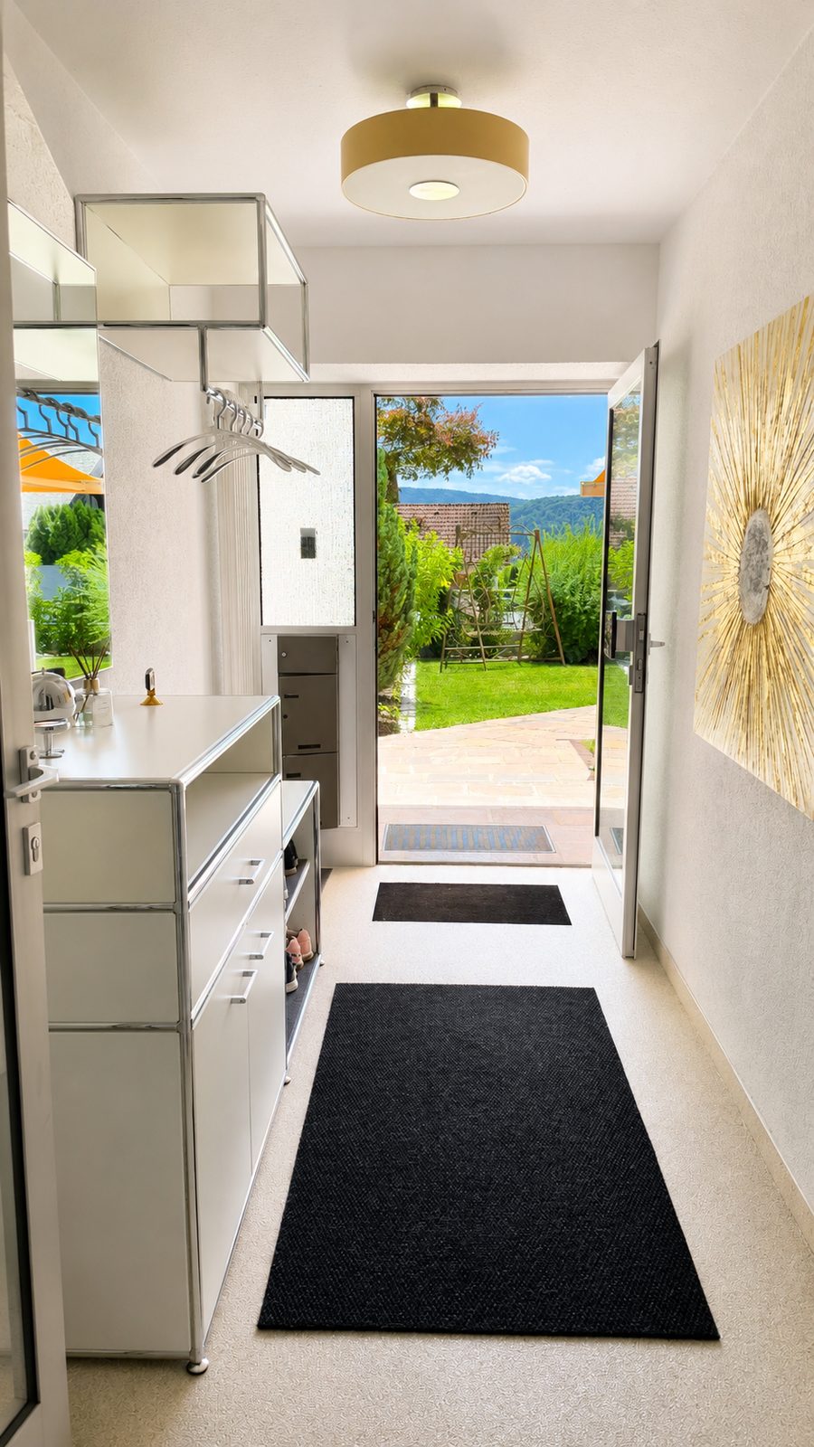 Eingangsdiele mit weissem Sideboard und Garderobe, offene Tuer mit Blick auf Garten, blauen Himmel und Berg mit Burgruine