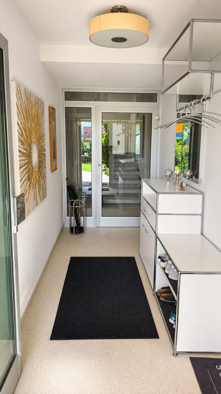 Eingangsdiele mit weissem modularem Sideboard, Garderobe mit Buegeln, Designleuchte, Sonnen-Wandbild und Glastuer mit Blick ins Treppenhaus
