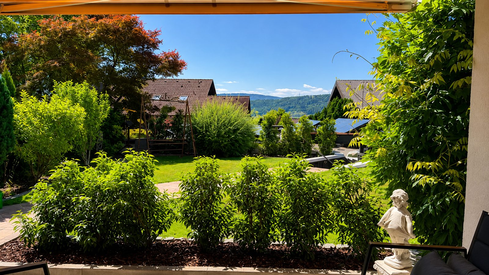 Gartenblick mit rotlaubigem Ahorn, Hecken, Rasen und Huegelpanorama mit Nachbarhaeusern unter blauem Himmel