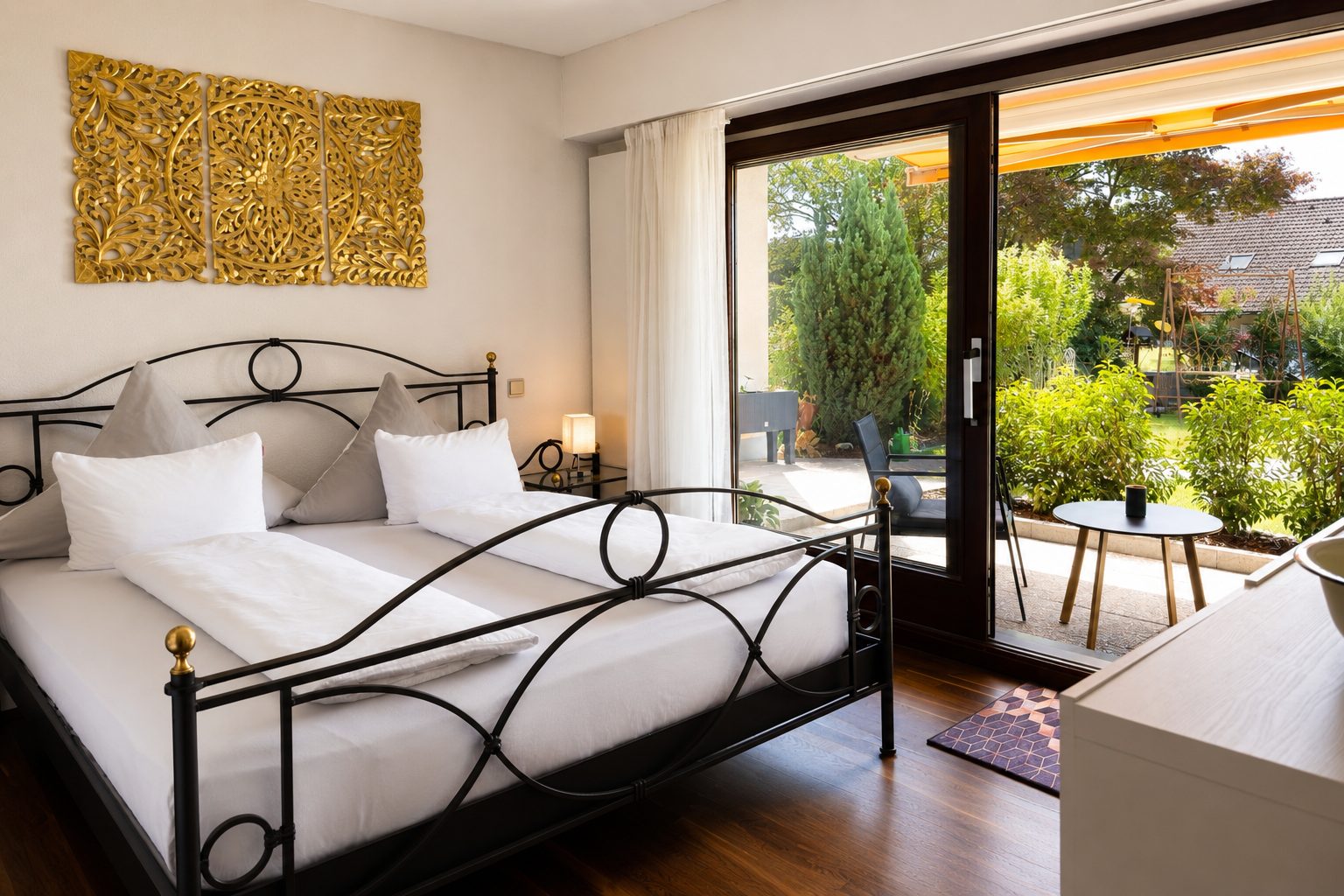 Schlafzimmer mit schwarzem Metall-Doppelbett, goldenem dreiteiligem Wandornament, Nachttischlampe und grosser Glasschiebetuer mit Blick in den Garten