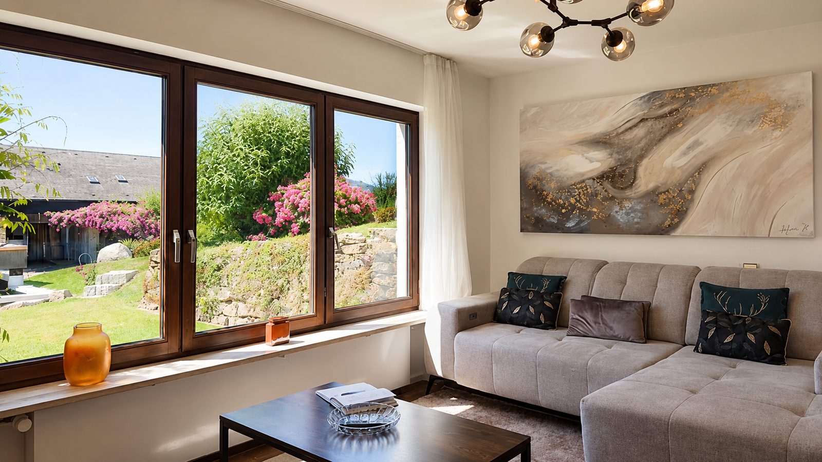 Wohnzimmer mit grauer Eckcouch, Designer-Kugelleuchte und Eckfenster mit Blick in den bluehenden Garten bei warmem Sonnenlicht