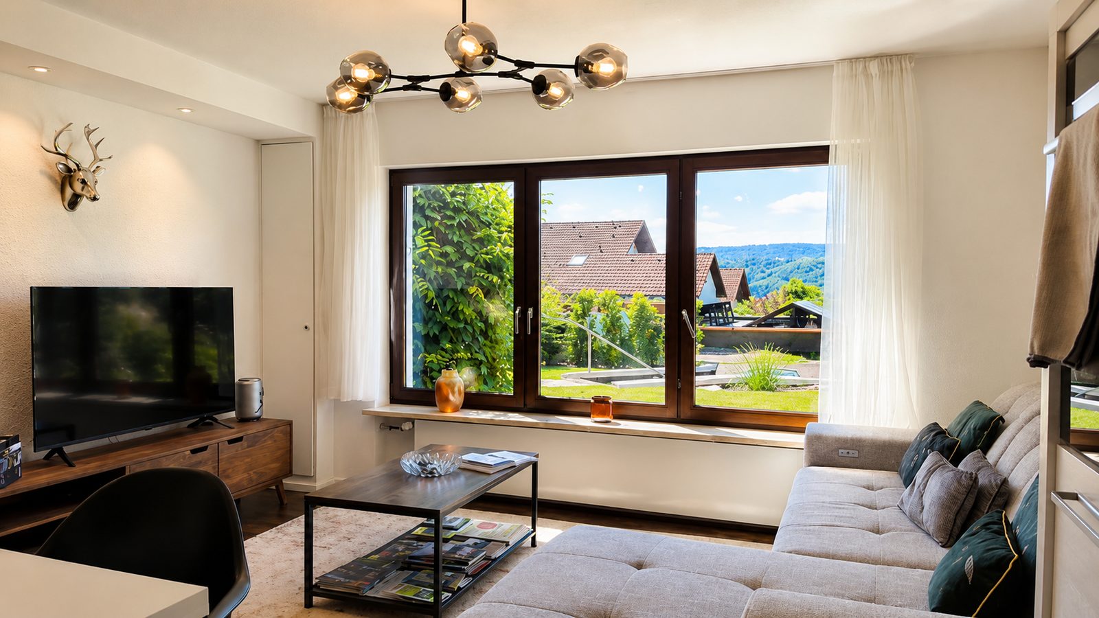 Wohnzimmer mit TV-Lowboard, Couch und grossem Panoramafenster mit Blick auf Haeuser und die Huegellandschaft