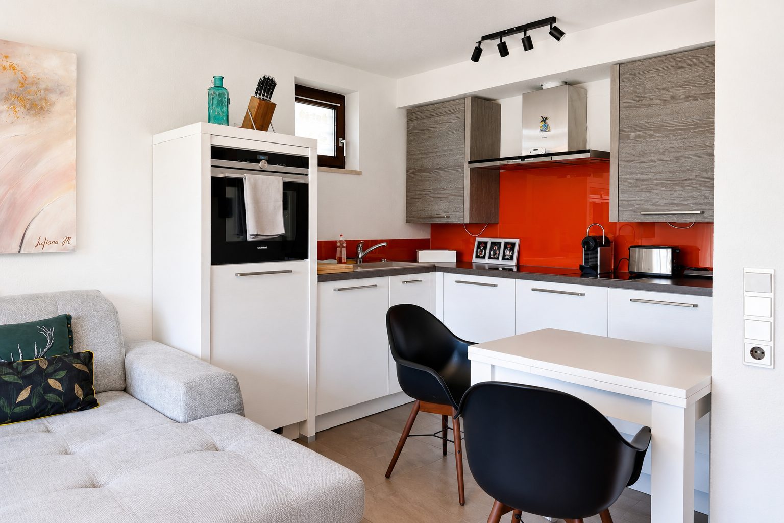 Offener Wohn- und Kuechenbereich mit Sofa, kleinem Esstisch mit schwarzen Schalenstuehlen und weisser Einbaukueche mit roter Glasrueckwand